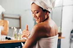 Woman applying lotion to her shoulder while smiling wearing a towel and a head wrap
